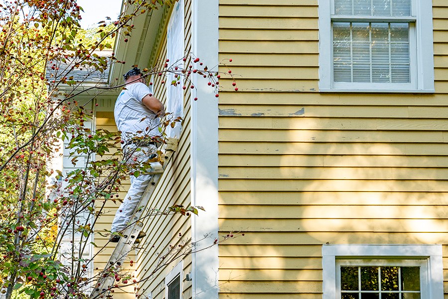 A painter on a ladder painting the exterior of a yellow house